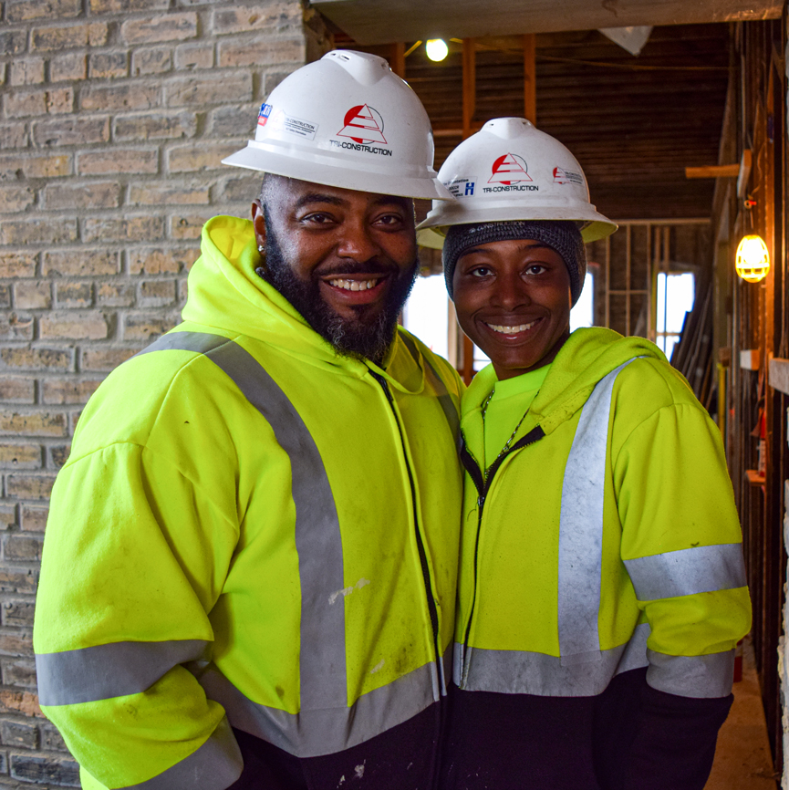 Two people smiling and posing for the camera wearing construction gear.